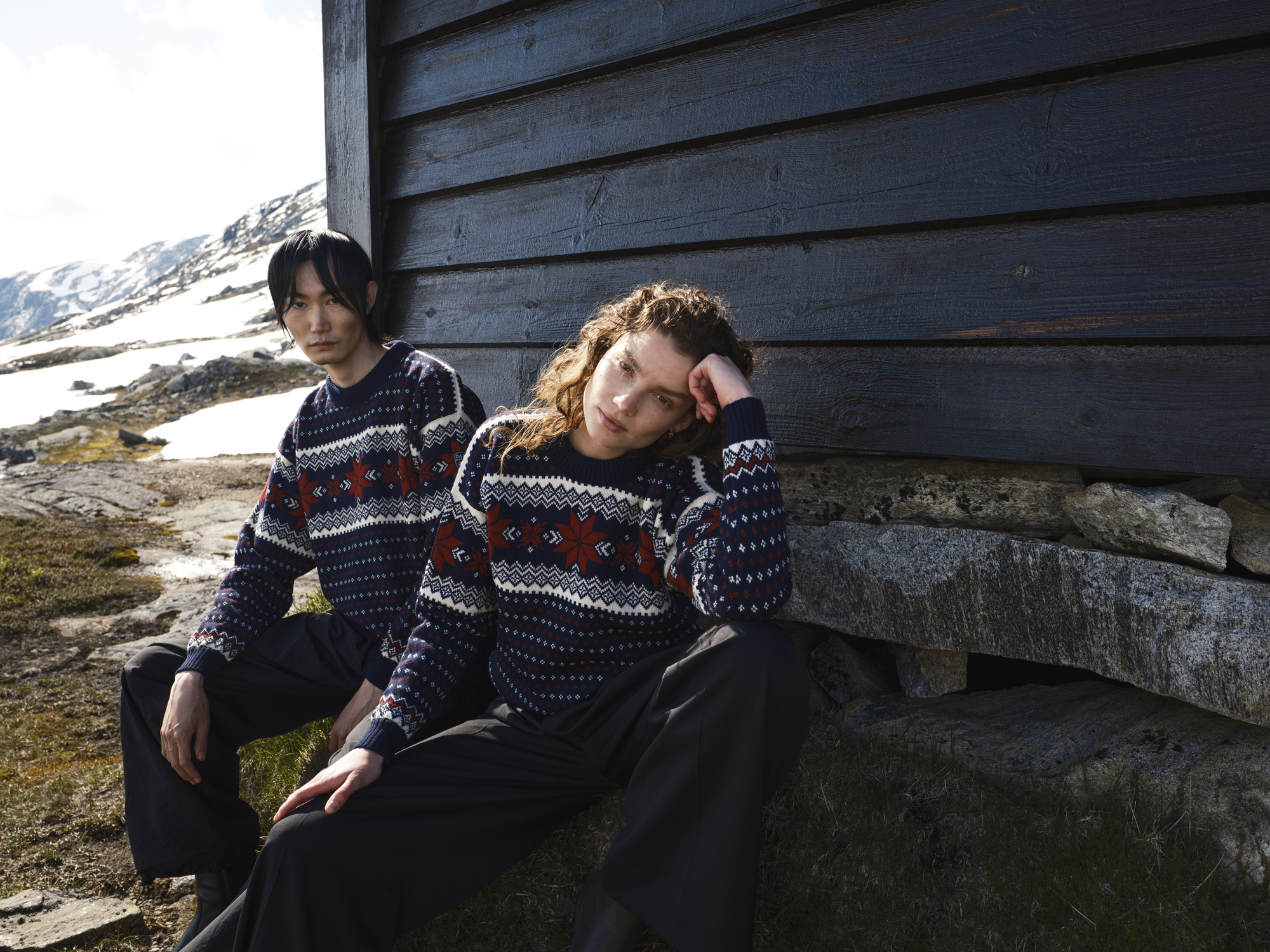 Male and female model sitting by a cabin in the Norwegian mountain wearing a Dale of Norway Unisex sweater